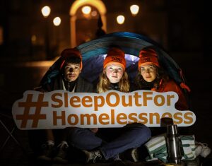 One young man and two young women sit in a tent in a low lit area with street lights with a sign that says "~Sleep Out for Homelessness"