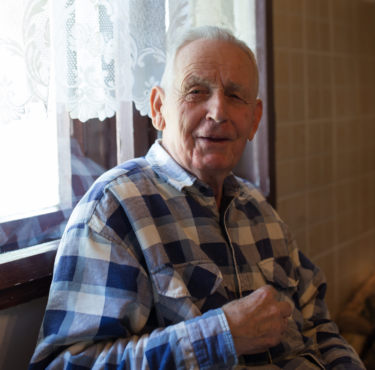 An elderly man with white thinning hair in a blue checked shirt smiles